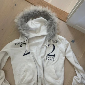  fur zipup  - Vit hoodiejacka från Hollister med grå fuskpäls runt huvan och tryck på bröstet. Jackan har dragkedja, fickor framtill och ribbade muddar. 