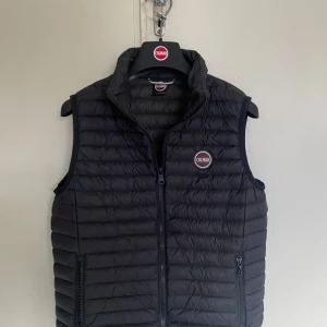 Svart dunväst från Colmar - Snygg svart dunväst från Colmar med quiltad design och dragkedja framtill. Västen har två praktiska fickor med dragkedja och en hög krage. Ikonisk Colmar-logga på bröstet ger en sportig vibe. Perfekt att slänga över en hoodie eller tröja. Priset är absolut diskuterbart👍👍