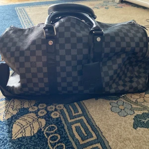 Louis Vuitton Damier Keepall resväska - Säljer en klassisk Louis Vuitton Keepall i svart och grått Damier-mönster. Väskan har dubbla handtag i svart skinn, silverfärgade metalldetaljer och dragkedja upptill. Perfekt storlek för weekendresor och riktigt snygg att bära med sig. Priset kan även diskuteras via dm