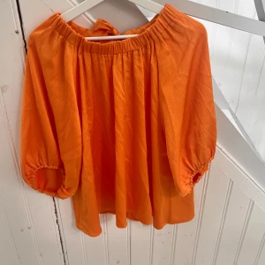 Orange blus från Stockh LM - Superfin orange blus från Stockh LM med vida trekvartsärmar och resår vid halsringningen. Blusen har en snygg knytning i nacken och är gjord i ett lätt, strukturerat tyg som ger en luftig känsla. Perfekt för dig som vill sticka ut med färg!