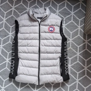 väst från Canada Goose - Snygg ljusgrå dunväst från Canada Goose med svart panel på sidorna och stort logotryck längs båda sidorna. Västen har broderad logga på bröstet, hög krage och dragkedja framtill. Perfekt för lager-på-lager och streetstyle. Det står S som storlek men jag använder M på allt annat och den passar fint på mig.
