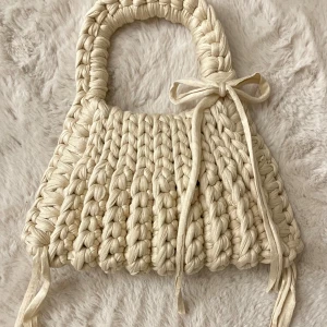Beige stickad handväska med rosett - Superfin handväska i beige stickat material med chunky flätad design och söt rosettdetalj framtill. Perfekt accessoar för att lyfta din outfit med en unik och trendig touch. Handtaget är också stickat och väskan har en mjuk, bohemisk vibe.
