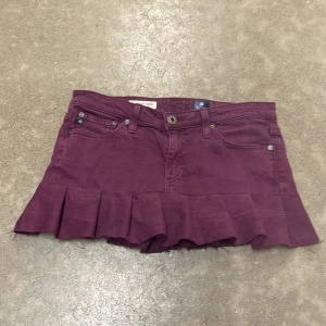 Mörklila Lågmidjad  jeanskjo - Mörklila kort jeanskjol med volangkant nertill. Storleken är 27 och ca XS och midijemåttet är ca75. 