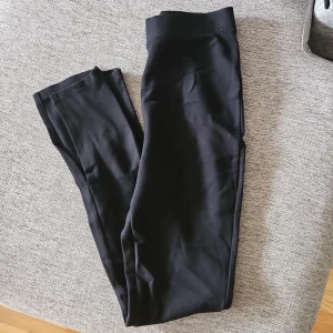 Svarta leggings med hög midja - Svarta leggings från NA-KD x Chloé B med hög midja och stretchigt material. Sitter tight och är perfekta för en chill vardagslook eller till träning. Mjuka och bekväma med enkel design och slits längst ner på benen.