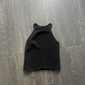 Svart stickad linnetopp från Lindex - Säljer en svart stickad linnetopp från Lindex i storlek S. Toppen har hög hals med knapp i nacken och är ärmlös. Snyggt virkad struktur med diskret mönster och croppad passform. 