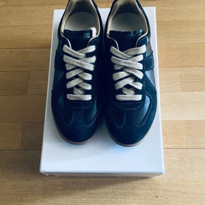 Maison Margiela Navy blue sneakers - Maison Margiela sneakers i svart skinn och mocka med vita snören och gummisula. Klassisk och stilren modell med rund tå och kontrasterande detaljer. Perfekta för dig som gillar minimalistisk design och vill ha något unikt på fötterna.