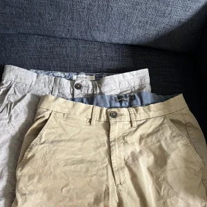 Beige och ljusgrå shorts Dressmann - Två par klassiska shorts från Dressmann, ett par i beige och ett i ljusgrått. Båda har normal passform, bälteshällor och sidfickor. Perfekta för varma dagar och enkla att matcha med t-shirt eller skjorta. Finns att hämta i Bromma