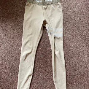Beige leggings från Stronger - Snygga beige leggings/tights från Stronger med Stretchigt material som sitter tight och bekvämt, perfekt för träning eller chill. Elastisk midja med logga runtom. Riktigt fräsch look!