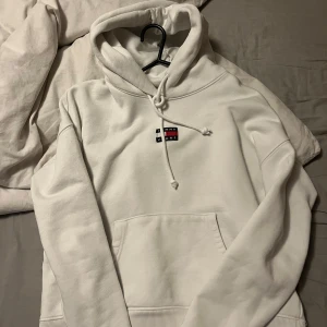 Vit hoodie från Tommy - Säljer en stilren vit hoodie från Tommy Jeans med klassisk logga broderad på bröstet. Tröjan har huva med snörning och en stor magficka framtill. Perfekt för dig som gillar enkel och snygg streetstyle.