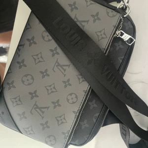 Louis Vuitton grå axelväska monogram - Snygg axelväska från Louis Vuitton i grått och svart med klassiskt monogrammönster. Väskan har bred svart axelrem med logga, silverfärgad dragkedja och är tillverkad i skinn. Perfekt för dig som vill ha en stilren och lyxig accessoar.