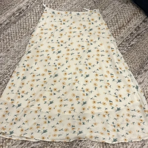 Blommig kjol från DAZY - Supersöt kjol från DAZY i ljusbeige med gult blommönster och gröna blad. Kjolen har en lätt A-linjeform och dragkedja i sidan. Storlek S