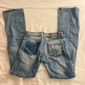 Bootcut jeans med strassdetaljer - Ljusblå bootcut jeans från Mc Lem med snygga slitningar och strass/diamant detaljer på bakfickorna. Klassisk femficksmodell med låg midja och dragkedja. Jeansen har en cool tvätt och markerade sömmar. Små gröna, blå och röda detaljer. Står ingen storlek men mått finns, skulle säga ungefär xs. (Vissa stenar har lossnat)