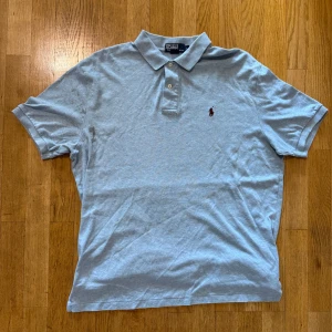 Grå pikétröja från Ralph Lauren - Klassisk grå pikétröja från Ralph Lauren med broderad logga på bröstet. Tröjan har krage, två knappar och korta ärmar. Perfekt för en clean och stilren look. Tillverkad i mjuk bomull som känns skön mot huden.