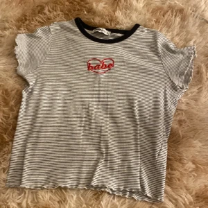 Randig topp med broderi från FB Sister - Superfin kortärmad topp från FB Sister i svartvitt randigt med broderad röd text 'babe' i ett hjärta på bröstet. Svart kant vid halsen och vågig finish vid ärmslut och nederkant. Perfekt för en chill och trendig look.