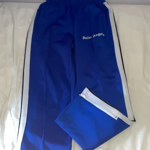 Blåa Palm Angels trackpants - Snygga blåa trackpants från Palm Angels med vita sidoränder och vit logga på låret. Byxorna har elastisk midja och dragkedja vid benslutet. Perfekta för en sportig och trendig look.