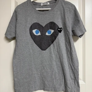 Grå Comme des Garçons Play t-shirt - Grå t-shirt från Comme des Garçons Play med klassiskt svart hjärta med ögon och liten hjärtdetalj på bröstet. T-shirten har rund hals, korta ärmar och är gjord i mjuk bomull. Perfekt för dig som gillar streetstyle och vill sticka ut med en ikonisk design.
