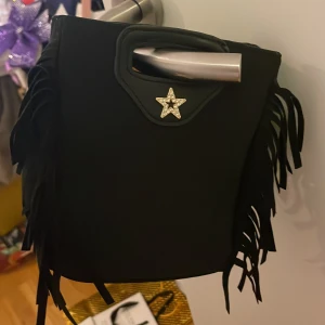 Svart fransväska från Gina Tricot - Cool svart handväska från Gina Tricot med fransar på sidorna och ett handtag upptill. Väskan har en dekorativ stjärna med strass framtill och är tillverkad i syntetmaterial. Perfekt för dig som vill sticka ut med en edgy accessoar.