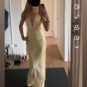 Ljusgul långklänning med djup v-ringning - Säljer en elegant ljusgul långklänning i satinliknande material. Klänningen har djup v-ringning framtill, smala axelband och öppen rygg med snörning. Sitter figurnära och ger en riktigt snygg siluett. Perfekt för dig som vill sticka ut!