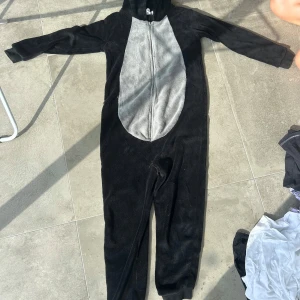 Svart och grå one-pice i mjukt material  - Mysig svart hoodie från Lazy One i fleece med grå mage och dragkedja framtill. Har huva och långa ärmar, perfekt för chill dagar hemma eller som rolig pyjamas. Supermjuk och skön att ha på sig! Har hål mellan benen som enkelt går att laga. Nypris 200, pris kan diskuteras