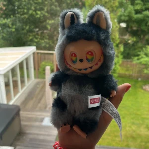 Labubu äkta / secret - Supergullig liten väska/nyckelring från Pop Mart i form av en fluffig figur med grå och svarta nyanser. Väskan har pälsliknande material, stora öron och regnbågsfärgade ögon. Perfekt att hänga på väskan eller använda som accessoar.
