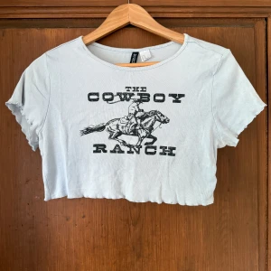 Vit croppad cowboy t-shirt - Vit croppad t-shirt från Chiquelle med svart cowboy-tryck och texten 'The Cowboy Ranch' framtill. T-shirten har korta ärmar och vågig kant nertill och vid ärmslut. Perfekt för dig som gillar western vibes 