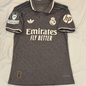 Real Madrid x Adidas grå matchtröja - Snygg grå Real Madrid fotbollströja från Adidas med broderade klubbmärken, HP-logga och Champions League 15-patch på ärmen. Tröjan har korta ärmar, krage med knappar och subtilt mönster i tyget. Denna tröjan är samma som spelarna använder(player version). Tröjan är från 2025 och är i extremt bra skick. Säljs billigt.