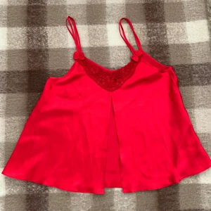 Röd satin linnetopp med spets - Supersöt röd linnetopp i satin med dubbla smala axelband och små rosetter. Toppen har v-ringning med broderad spets och blommiga detaljer framtill. Lätt utsvängd passform som ger en luftig känsla. Perfekt för dig som vill sticka ut med en färgstark look.