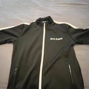Palm Angels svart track jacket - Svart track jacket från Palm Angels med vita ränder längs ärmarna och vit dragkedja. Jackan har hög krage, två fickor med dragkedja och logga på bröstet. Perfekt för en sportig och clean streetstyle.
