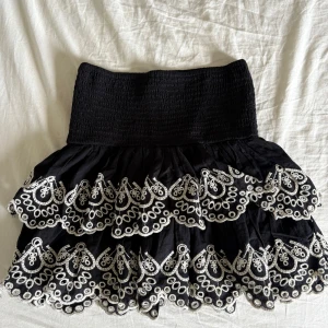 Svart volangkjol med broderi från Lindex - Supersöt svart kortkjol från Lindex med bred smock i midjan och flera lager volanger. Kjolen har vit broderad spetsdetalj längs volangerna