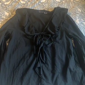 Svart blus med volang och knyt från 157 - Svart blus från 157 i storlek XS sitter som en S, med långa ärmar, volangdetaljer längs ringningen och snygg knytning framtill. Tunt och lätt material, väldigt luftig men ändå lite varm, använd 1 gång 