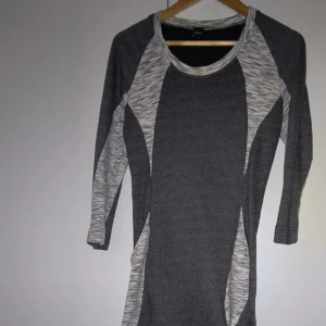 Grå och vit trikåklänning med mönster - Snygg grå klänning med vita, melerade paneler och trekvartsärm. Figurnära passform och sportig känsla, perfekt för dig som gillar stilrena och bekväma plagg. Mjuk trikå som känns skön mot huden.