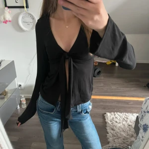 Svart långärmad knytblus XS - Svart långärmad topp med djup v-ringning och knyt framtill. Modellen har vida ärmar och croppad passform som ger en trendig look. Perfekt att styla med jeans eller kjol för en cool vibe.