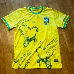 Brasilien Nike gul fotbollströja fågel - Gul Brasilien fotbollströja från Nike med gröna detaljer och tryck av två stiliserade fåglar på framsidan. Tröjan har korta ärmar med gröna och blå muddar samt CBF-emblem på bröstet. Tillverkad i lätt och ventilerande material, perfekt för match eller träning.
