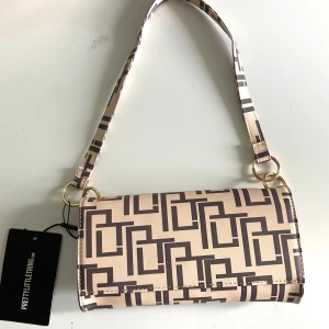 Beige axelväska från PrettyLittleThing - Snygg beige axelväska med grafiskt mönster i brunt från PrettyLittleThing. Väskan har guldfärgade metalldetaljer och ett stilrent, rektangulärt format. Perfekt för att lyfta din outfit med en trendig touch.