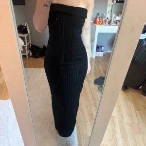 Svart axelbandslös långklänning - Säljer en svart axelbandslös långklänning med smala vita ränder. Klänningen är figurnära och har en slits därbak. En väldigt fin klänning som aldrig kommit till användning 