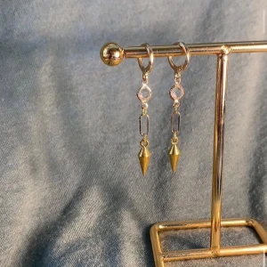 Guldiga örhängen med spetsig detalj - Snygga guldiga örhängen med hängande design. De har en rund ögla, en liten transparent pärldetalj och en avlång länk som avslutas med en spetsig konformad guldpendel. Perfekta för att ge din look en cool edge.