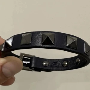 Valentino svart läderarmband med nitar - Svart läderarmband från Valentino Garavani med ikoniska pyramidformade silverfärgade nitar. Justerbart spänne i metall och stilren design som ger en edgy vibe. Perfekt accessoar för att lyfta din outfit med en lyxig touch.
