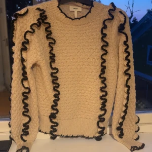 Stickad beige tröja med svarta volanger - Beige stickad tröja från Object med svarta volangdetaljer längs ärmar och sidor. Tröjan har rund halsringning, ribbade muddar och en cool struktur i stickningen. Perfekt för dig som vill ha något unikt och trendigt i garderoben.