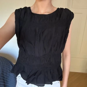 Svart blus - Säljer en svart ärmlös blus med volangdetalj nedtill och rynkad effekt. 