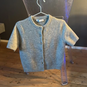 Grå kortärmad kofta från Zara - Säljer en grå kortärmad kofta från Zara med rund halsringning och knappar framtill. Tröjan är stickad och har en klassisk, enkel design som passar till mycket. Perfekt för lager-på-lager-stil. Koftan saknar dock en knapp nedtill på tröjan men kan lätt ersättas med en ny knapp!