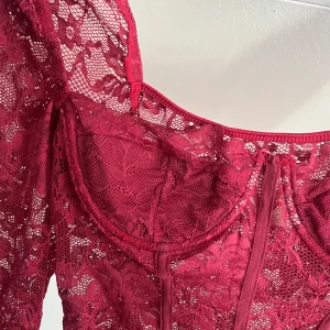 Vinröd spetsbody med lång ärm - Säljer en vinröd korsettopp i spets med långa ärmar och markerade kupor. Toppen har en figurnära passform och vacker blommig spetsdetalj över hela plagget. Perfekt för dig som vill sticka ut med en elegant och trendig look.