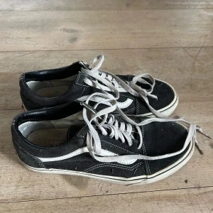 Svarta och vita sneakers från Vans - Klassiska svarta och vita Vans sneakers med vit sula och vit sidestripe. Skorna har snörning och en platt gummisula med det karakteristiska rutmönstret undertill. Perfekta för en avslappnad streetstil.