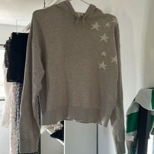Beige hoodie med stjärnor - Säljer en beige hoodie med vita stjärnor på ena axeln. Tröjan har huva och långa ärmar, samt en croppad passform. Perfekt för en avslappnad stil.