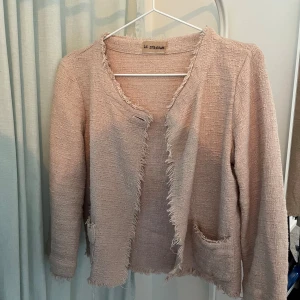 Ljusrosa cardigan jacka från Le Streghe - Säljer en ljusrosa cardigan jacka från Le Streghe med råa kanter och två fickor framtill. Jackan har en öppen design utan knäppning och är tillverkad i ett strukturerat tyg som ger en exklusiv känsla.