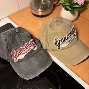 Två kepsar Grancanaria - Två kepsar i vintage-stil från Grancanaria Original, en i grått med röd och vit broderad text och en i beige med blå och vit broderad text. Båda har böjd skärm och slitna detaljer för en avslappnad look.