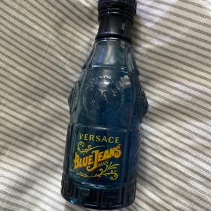 Versace Blue Jeans Man parfym - Säljer en ikonisk herrparfym, Blue Jeans Man från Versace. Flaskan är blå och har en unik, detaljerad design med gul text. Parfymen har en fräsch och maskulin doft, perfekt för dig som gillar klassiska och stilrena dofter.