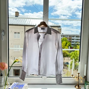 Vit skjorta med grå detaljer från NA-KD - Säljer en stilren vit skjorta. Skjortan har klassisk krage, knäppning framtill och långa ärmar. Den är väl använd och har sina fläckar, inget som vanish inte kan ta bort.