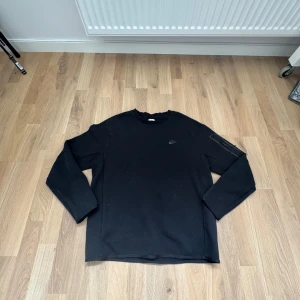 Svart långärmad tröja från Nike (L) - Säljer en svart långärmad tröja från Nike med rund hals och diskret Nike-logga på bröstet. Tröjan har en ficka med dragkedja på ena ärmen och är tillverkad i mjukt material. Perfekt för en sportig och avslappnad stil.