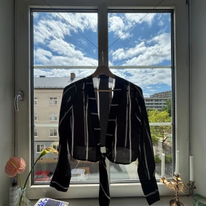 Svart randig knytblus från PrettyLittleThing - Säljer en svart blus med vita ränder från PrettyLittleThing. Blusen har långa ärmar och knytning framtill, vilket ger en snygg och trendig look. Perfekt att styla med höga byxor eller kjol.