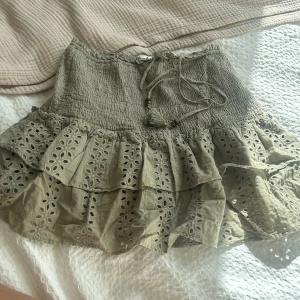 Grön volangkjol med broderade detaljer - Supersöt beige kjol med volanger och broderade blommönster. Kjolen har smock i midjan och knytband med tofsar framtill. Perfekt för en somrig och avslappnad look.